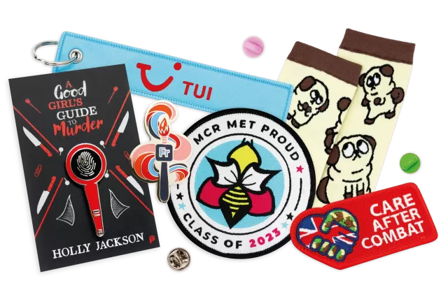 A collection of custom merch create by Made by Cooper for their clients. A school woven patch, a military embroidered patch, a Good Girl's Guide to Murder, a TUI suitcase keyring, a pair of Dead Endia custom socks, and an Adobe branded hard enamel pin.