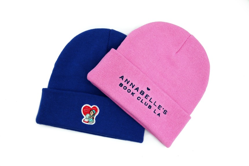A pink and blue custom beanie. The blue hat features a love heart logo while the pink hat has