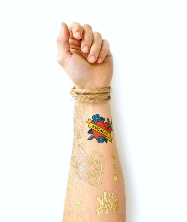 Forearm covered with colourful and gold custom temporary tattoos, including heart and hashtag motifs, showcasing mixed metallic and printed tattoo designs.
