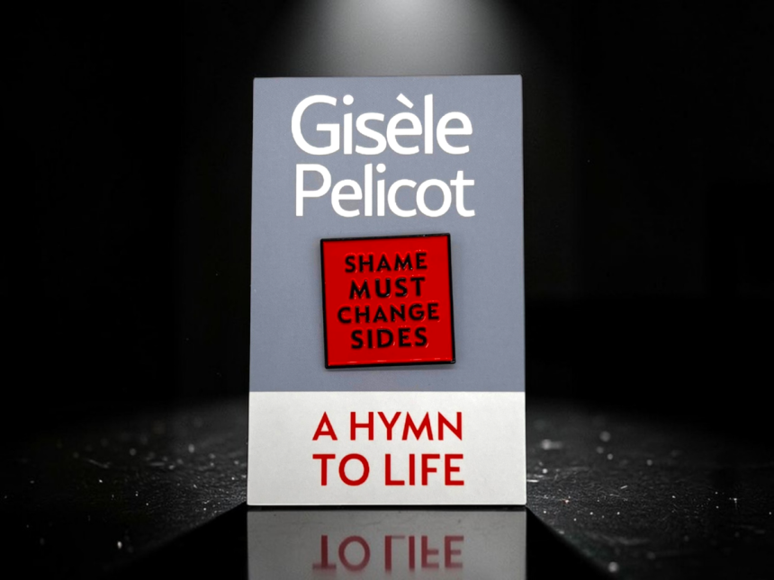 A Hymn to Life by Gisele Pelicot standing under dramatic spotlight lighting, featuring the red “Shame Must Change Sides” hard enamel pin badge centred on the grey book cover, with a reflective black surface beneath creating a subtle mirrored effect.