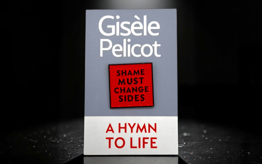A Hymn to Life by Gisele Pelicot standing under dramatic spotlight lighting, featuring the red “Shame Must Change Sides” hard enamel pin badge centred on the grey book cover, with a reflective black surface beneath creating a subtle mirrored effect.