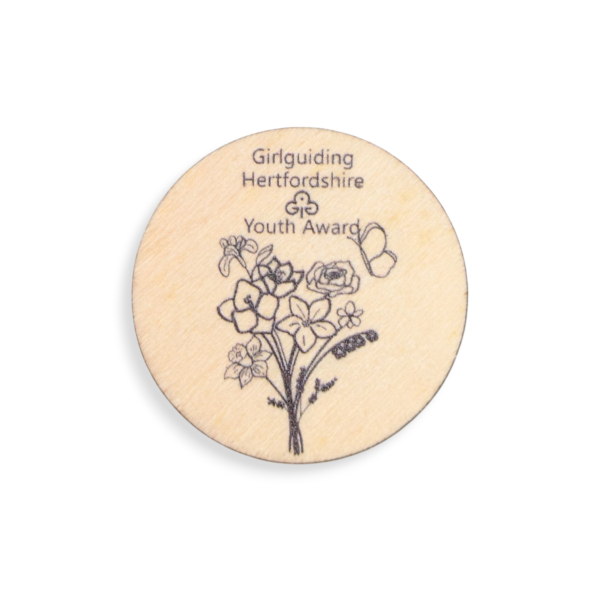 A circular wooden badge with a floral design and the words Girlguiding Hertfordshire Youth Award etched into it.